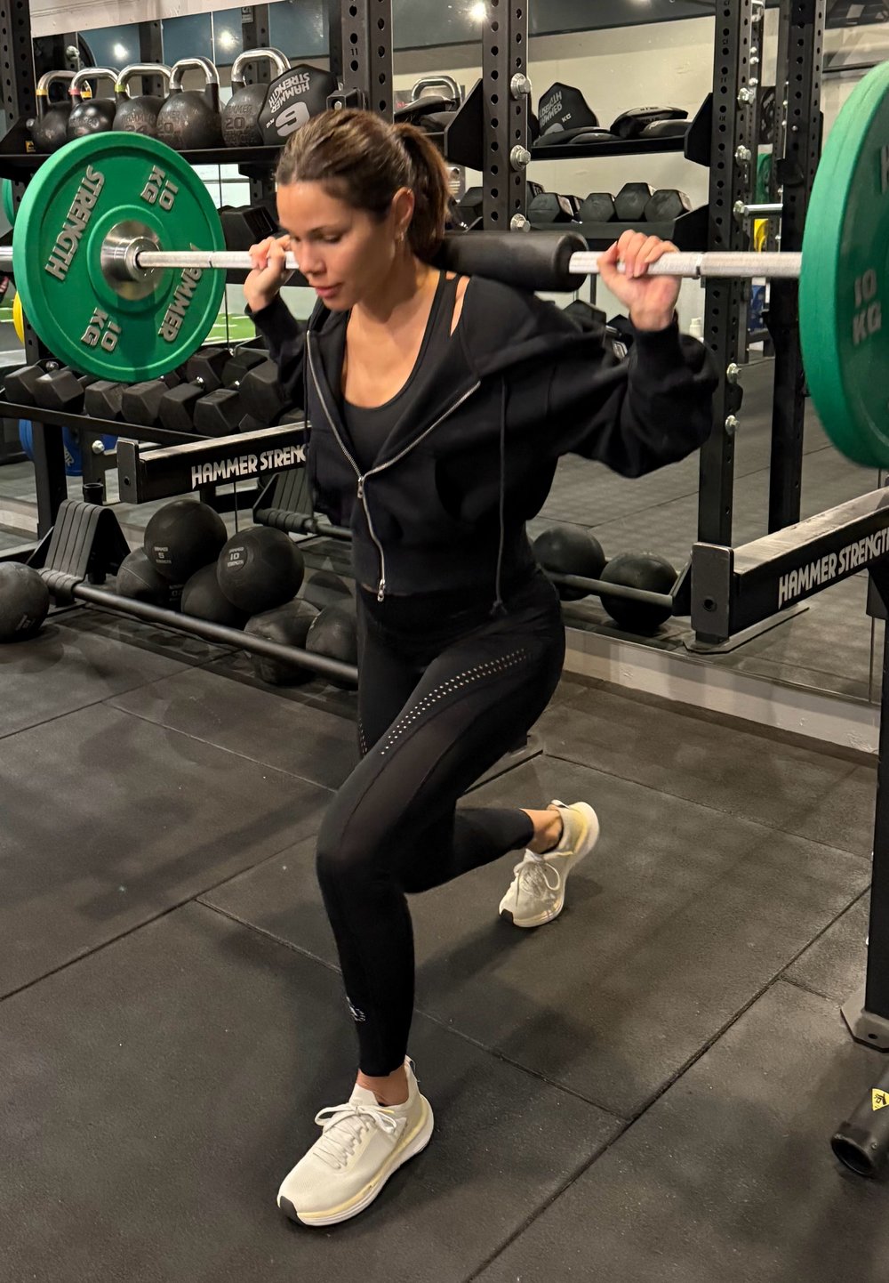 Athlète effectuant une fente avec une barre ; portant un sweat à capuche noir zippé, un legging noir avec un motif latéral et des baskets claires. Des poids verts sont utilisés.