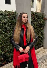 Mustast nahast mantel, punane kampsun ja narmastega punane sall. Käes väike punane käekott. Taustal tihe roheline lehestik ja hall arhitektuur.