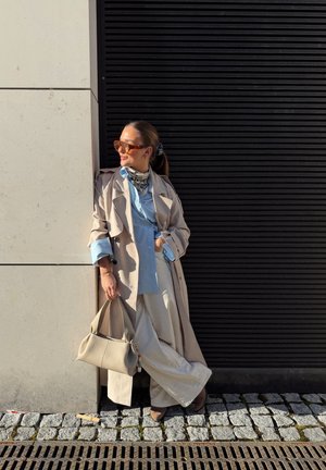 Mujer con gabardina beige, camisa azul claro, pantalones blancos, gafas de sol y pañuelo se apoya contra una pared sosteniendo un bolso beige en una calle adoquinada.