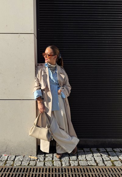 Mujer con gabardina beige, camisa azul claro, pantalones blancos, gafas de sol y pañuelo se apoya contra una pared sosteniendo un bolso beige en una calle adoquinada.