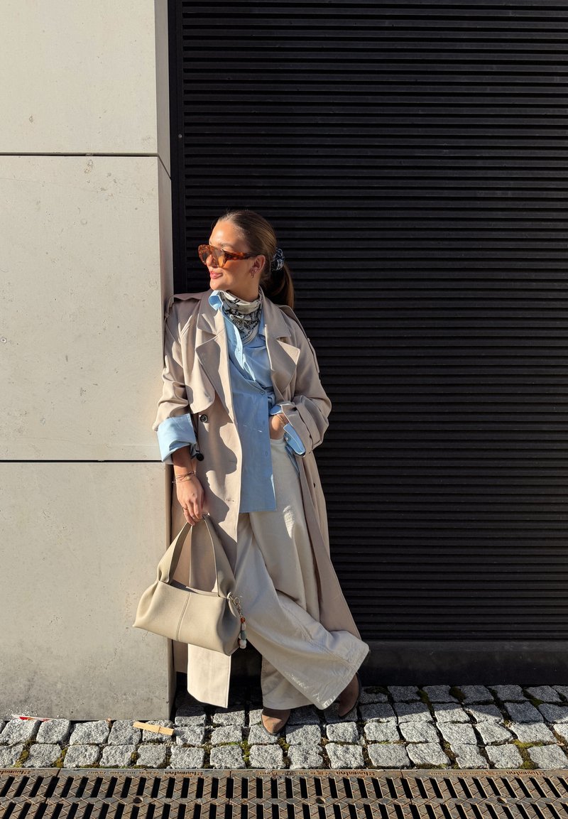Femme en trench-coat beige, chemise bleu clair, pantalon blanc, lunettes de soleil et écharpe, appuyée contre un mur et tenant un sac à main beige dans une rue pavée.