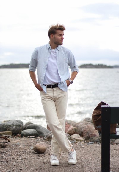 Camisa de botón a rayas azul claro sobre una camiseta blanca, combinada con pantalones beiges y zapatillas blancas; de pie junto a una ribera rocosa.