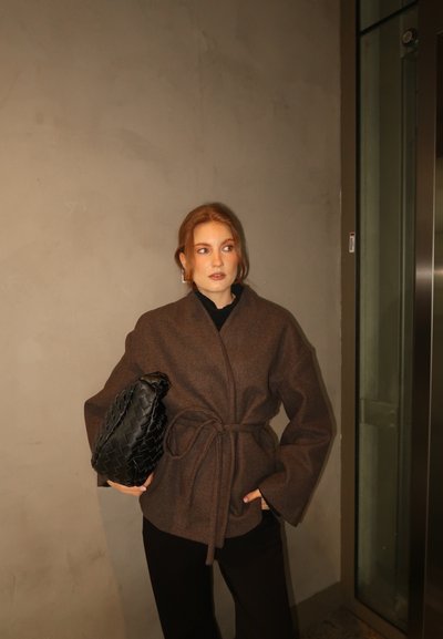 Mujer con cabello rojo que lleva un abrigo marrón con cinturón y pantalones negros, sosteniendo un gran bolso de mano negro tejido, parada cerca de un ascensor.