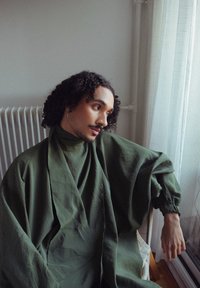 Oversized olijfgroene top met hoge kraag, gestructureerde stof en wijde mouwen. Persoon in een ontspannen pose binnenshuis.