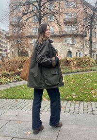 Ciemnozielona wodoodporna kurtka z dużą przednią kieszenią, w połączeniu z ciemnymi dzwonowatymi dżinsami. Na jednym ramieniu zawieszona jest brązowa torba.