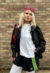 Svart skinnjakke over en hvit t-skjorte med en liten logo, svart skjørt med grønne striper og en rød rutete cap. Nøkkelring synlig.