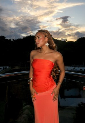Mujer con el cabello trenzado que lleva un vestido naranja sin tirantes, pendientes de flores y pulseras, de pie en un balcón al atardecer con colinas al fondo.