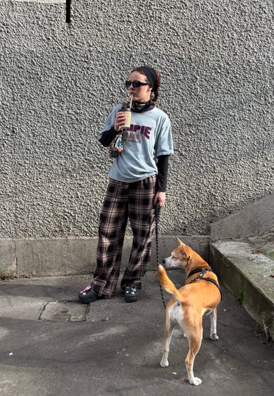 Joven mujer que lleva pantalones de cuadros y gafas de sol bebe de una taza mientras sostiene a un perro con correa frente a una pared gris con textura.
