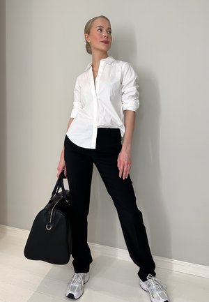 Camisa blanca de botones, pantalones negros, zapatillas grises y un bolso negro. Tela suave y ajuste informal, de pie contra una pared clara.