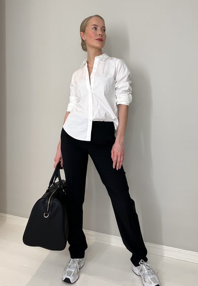 Camisa blanca de botones, pantalones negros, zapatillas grises y un bolso negro. Tela suave y ajuste informal, de pie contra una pared clara.