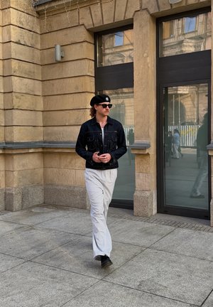 Hombre con chaqueta vaquera negra, pantalones blancos a rayas finas, boina negra y gafas de sol caminando por una acera urbana cerca de un edificio de piedra beige.