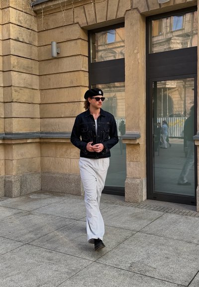 Hombre con chaqueta vaquera negra, pantalones blancos a rayas finas, boina negra y gafas de sol caminando por una acera urbana cerca de un edificio de piedra beige.