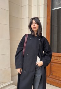 Čierny vlnený kabát s dvojradovým zapínaním, veľkými gombíkmi a širokými rukávmi. Kombinovaný s tmavosivými nohavicami a zapletenou bordovou kabelkou.