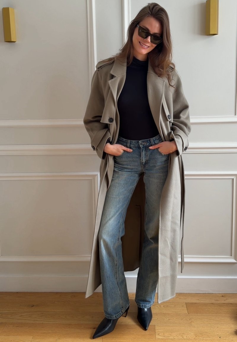 Femme portant un long manteau beige, un haut noir, un jean bleu, des bottes noires pointues et des lunettes de soleil, debout à l'intérieur contre un mur blanc à panneaux.