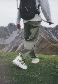 Sneaker mit einer massiven weißen Sohle, neon-gelben Akzenten und Mesh-Details. Getragen mit olivgrünen Hosen und einem schwarzen Rucksack auf Gras.