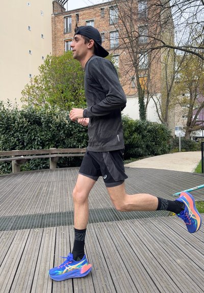 Joven corriendo al aire libre con ropa deportiva negra y zapatillas de correr de colores en un camino de madera cerca de arbustos verdes y edificios.