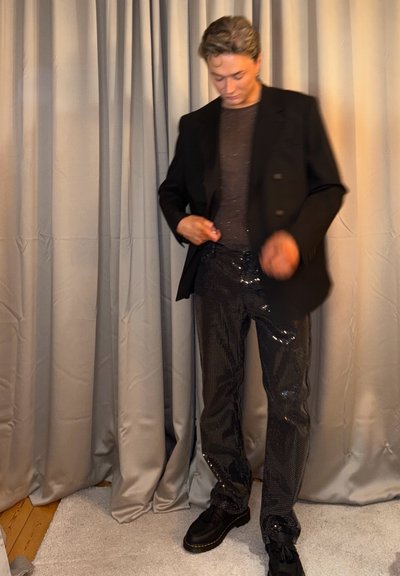 Blazer negro ajustado sobre una blusa oscura y brillante y pantalones cubiertos de lentejuelas. Zapatos negros con acentos texturizados. Cortinas grises al fondo.