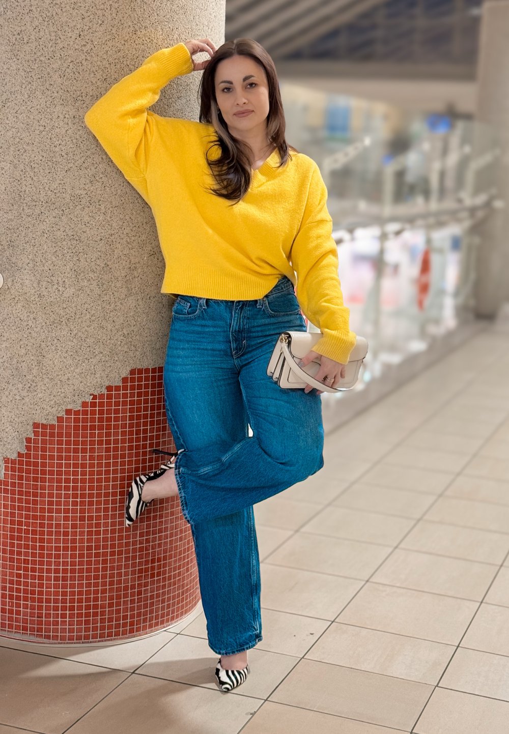 Femme en pull jaune et jean bleu s'appuie contre un pilier, tenant un sac beige, avec une jambe pliée, à l'intérieur sur un sol carrelé.