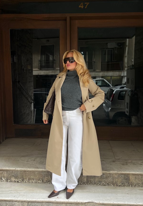 Mujer con largo cabello rubio, con gafas de sol, abrigo beige, jersey de cuello alto gris, pantalones blancos y tacones oscuros, parada en las escaleras fuera de un edificio.