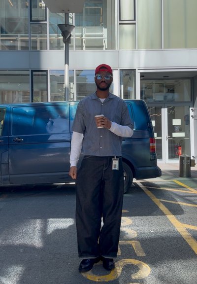 Camisa de rayas de mangas cortas con mangas largas blancas debajo, pantalones oscuros y holgados, gafas de sol, gorra roja, sosteniendo un vaso, de pie junto a una furgoneta azul.