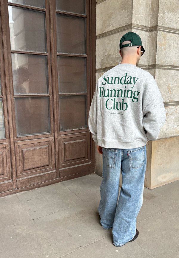 Mężczyzna w szarej bluzie z napisem „Sunday Running Club”, w niebieskich dżinsach, ciemnozielonej czapce, stoi przy drewnianych drzwiach i kamiennej ścianie.