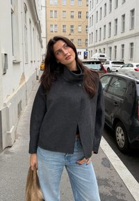 Donkergrijze, kraagloze jas met een iets oversized ontwerp, textuurstof en knoopdetail, gecombineerd met lichtdenim jeans en een tan tas.