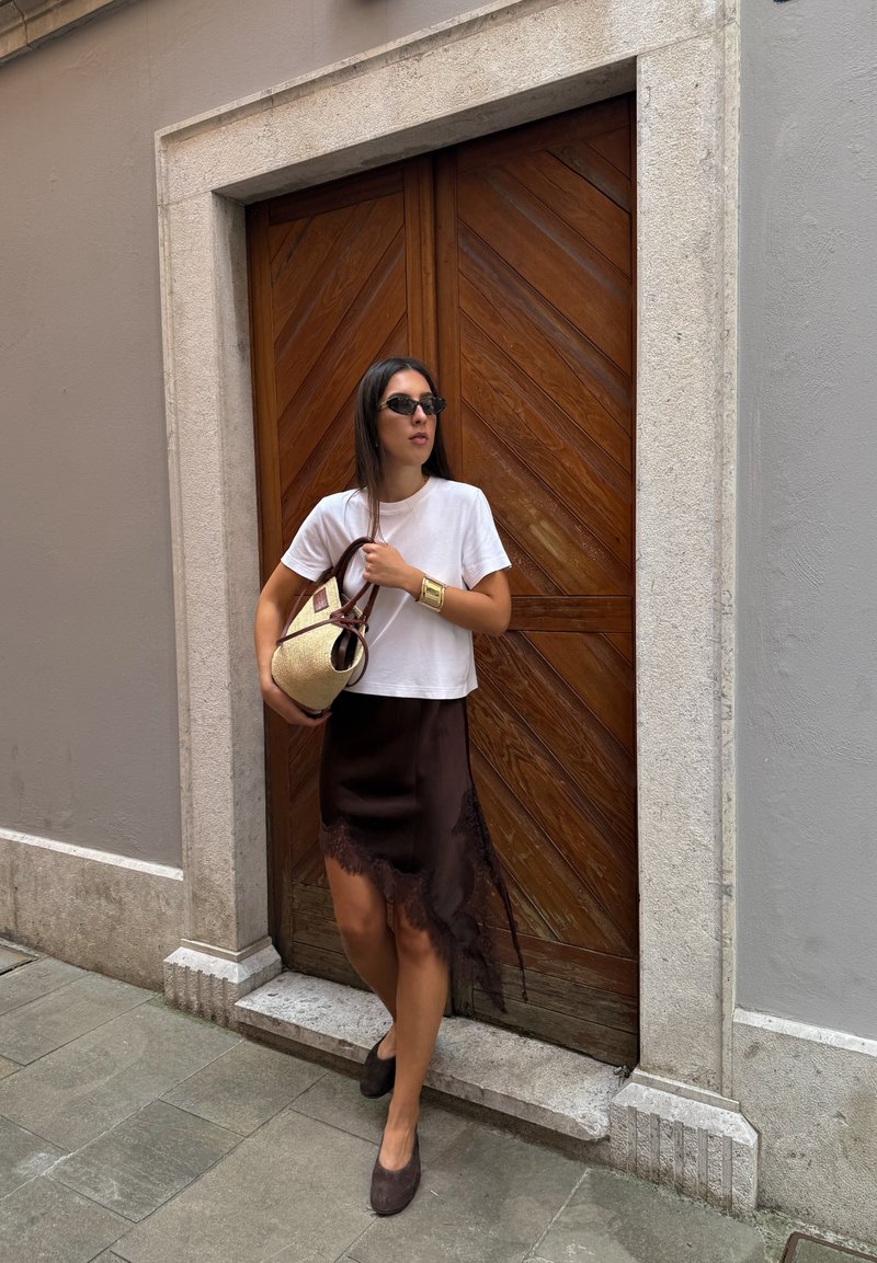 Camiseta branca cortada, saia de cetim castanha com detalhe de renda, óculos de sol e uma bolsa de palha com alças escuras, em pé ao lado de uma porta de madeira.
