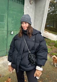 Černá péřová bunda s vysokým límcem a kapucí, doplněná o hnědé rukavice bez prstů. Šedá pletená čepice a černá kabelka přes rameno s řetízkem.