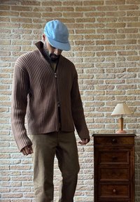 Suéter marrón de canalé con cremallera y cuello alto, combinado con pantalones color arena. Una gorra azul clara completa el conjunto, junto a una mesa lateral de madera con lámpara.