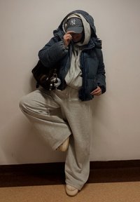 Jachetă matlasată bleumarin purtată peste un hanorac gri, asortată cu pantaloni sport gri și pantofi bej. O geantă închisă la culoare și o șapcă cu logo completează ținuta.