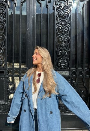 Trinchera de denin de gran tamaño en azul claro con botones negros, superpuesta sobre un suéter blanco, al aire libre frente a un fondo de reja de hierro forjado negra y ornamental.