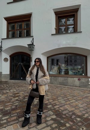Mujer con chaqueta de piel, pantalones negros y botas, de pie en una calle empedrada frente a un edificio blanco con ventanas de madera y puertas arqueadas.