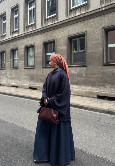 Mujer con un pañuelo rosa en la cabeza y una capa azul marino, sosteniendo un bolso marrón, parada en una calle de la ciudad frente a un edificio.
