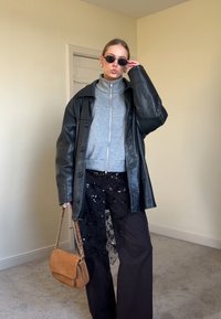 Femme portant une veste en cuir noire oversize, un sweat zippé gris, un pantalon large noir avec un revêtement en mesh à sequins, des lunettes de soleil, et tenant un sac à main beige.