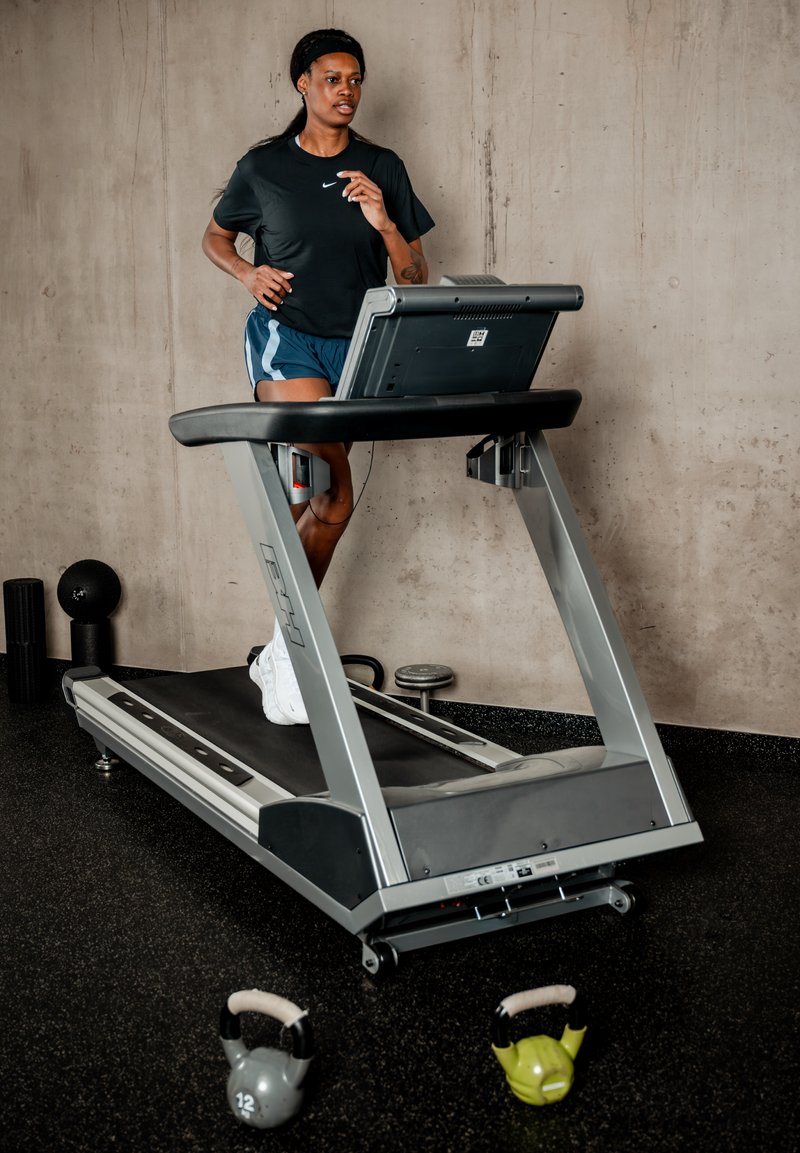 Femme portant un t-shirt noir et un short bleu courant sur un tapis de course dans une salle de sport avec des kettlebells et des poids à proximité.