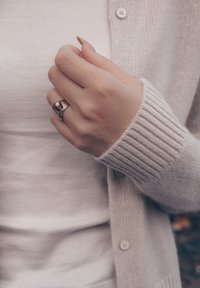 Srebrny pierścionek o trójkątnym wzorze, noszony na dłoni z zadbanymi paznokciami. Jasnobeżowy sweter zapinany na guziki z prążkowanymi mankietami, nałożony na biały top.