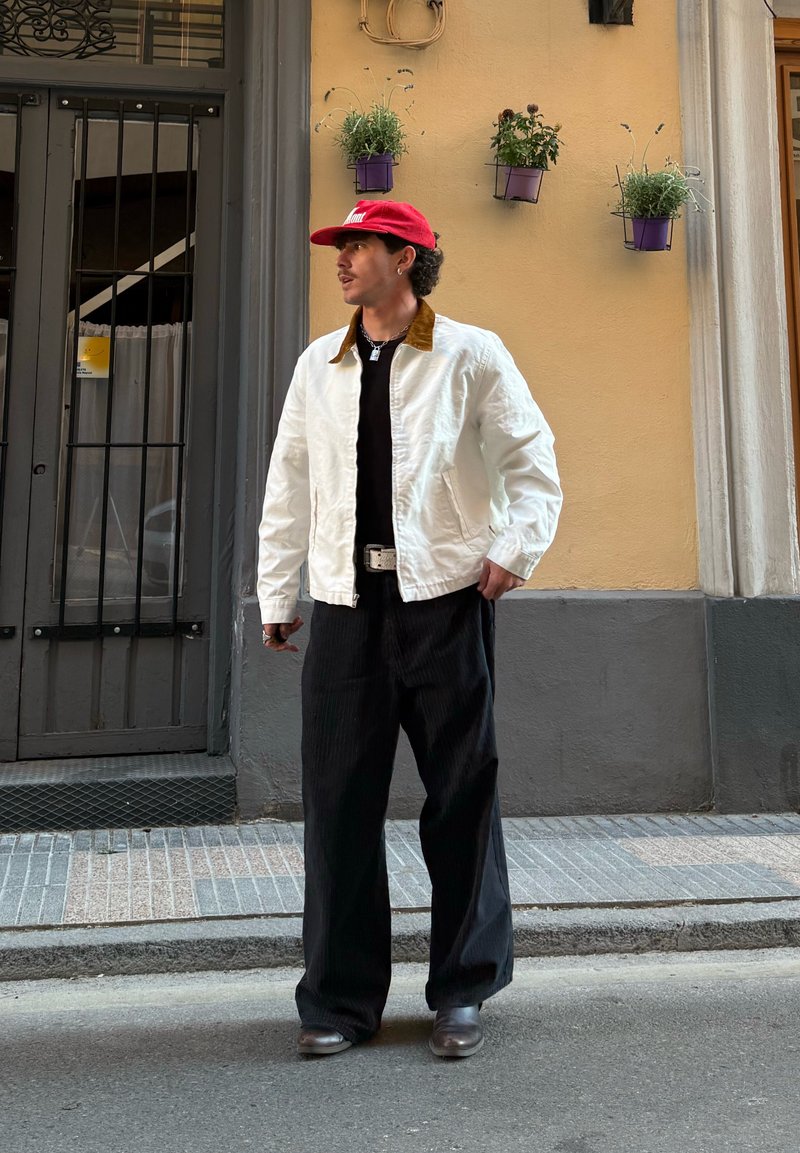 Jeune homme portant une veste blanche et une casquette rouge, debout dans une rue en ville près d'un bâtiment avec des pots de fleurs suspendus.