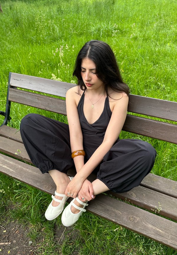 Jeune femme aux cheveux foncés portant une combinaison noire et des chaussures blanches, assise en tailleur sur un banc en bois dans un parc avec un fond d'herbe verte.