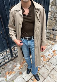 Beige színű trench coat, barna henley póló, kék farmernadrág, fekete öv, fekete cipők. A trench coat gombokkal ellátott és enyhén széles reverssel rendelkezik.
