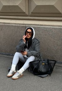 Femme en manteau gris, sweat à capuche blanc et leggings assise sur le trottoir contre un mur de pierre, tenant un téléphone, avec un grand sac à main noir à côté d'elle.