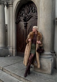 Manteau en fausse fourrure marron clair, pull texturé, mini-jupe verte, collants résille et bottes marron montantes jusqu'au genou. Sac à main marron et porte ornée en arrière-plan.