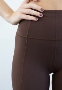 Leggings castanhos de cintura alta feitos de tecido elástico, com detalhes de costura visíveis e uma textura suave. Mão colocada no quadril.