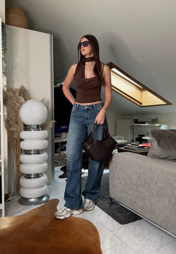 Junge Frau mit Sonnenbrille, braunem Neckholder-Top, blauen Jeans, grauen Sneakers, die eine schwarze Handtasche in einem modernen Dachbodenwohnzimmer hält.