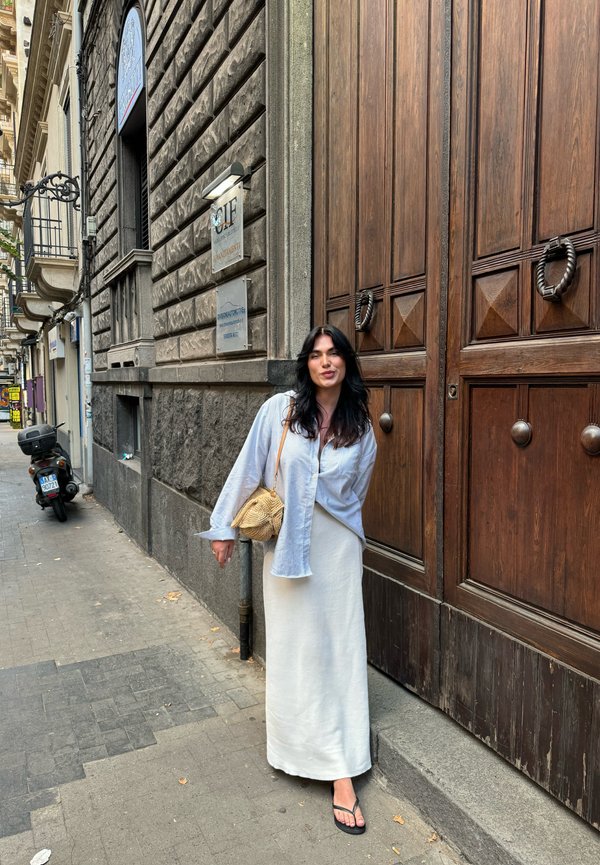 Cămașă bleu deschis, cu nasturi; fustă albă, texturată, până la gleznă; geantă din paie; flip-flops negre; uși din lemn în fundal.
