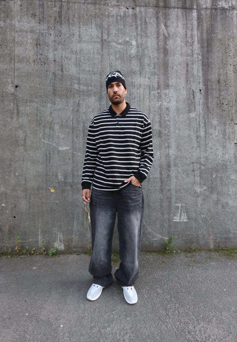 Polo rayé noir et blanc à manches longues, jean denim gris ample et baskets blanches. Porte un bonnet avec un logo, se tenant contre un mur en béton.