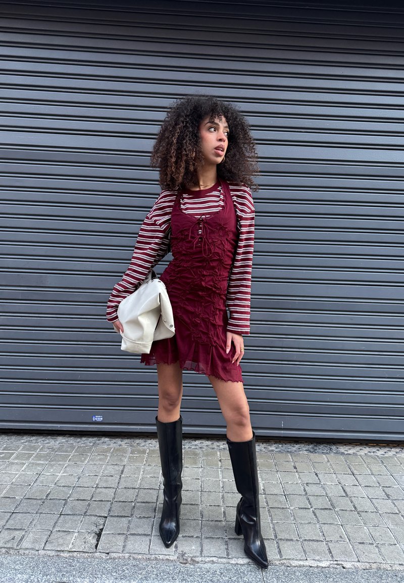Femme aux cheveux bouclés portant une robe bordeaux par-dessus un chemisier à manches longues rayé, des bottes noires jusqu'aux genoux, tenant un sac blanc, debout dans une rue.