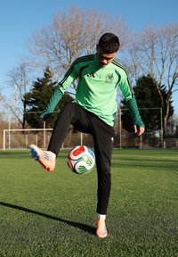 Jeune joueur de football masculin portant un maillot vert du Mexique jonglant avec un ballon multicolore sur un terrain herbeux, avec des buts et des arbres en arrière-plan.