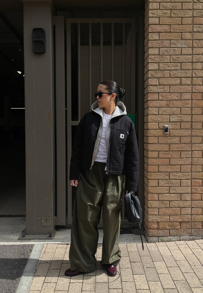 Frau mit Sonnenbrille, schwarzer Jacke, weißem Hemd und weitem olivgrünen Hose steht an einer Ziegelmauer und hält eine schwarze Handtasche.