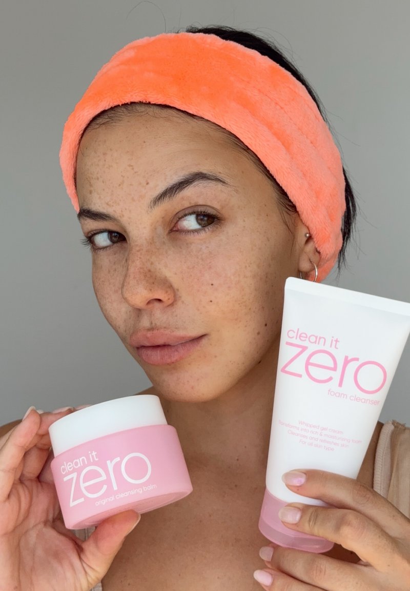 Femme avec un bandeau corail tenant près de son visage des produits de soin Clean It Zero baume démaquillant et nettoyant moussant roses.