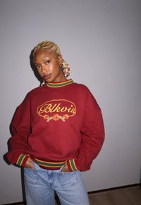 Sweatshirt marrom com "Blkvis" bordado, rosas amarelas e colarinho listrado em verde, vermelho e amarelo. Combinado com jeans azuis claros.
